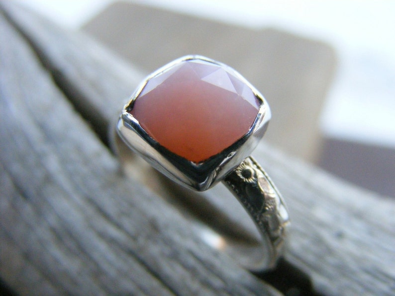 May include: A silver ring with a square, pink gemstone set in a bezel setting. The band is decorated with a floral design.