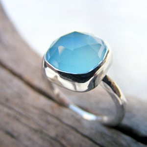 Puede incluir: Anillo de plata con una gema grande, de corte cuadrado, de color azul claro. La gema est&aacute; engastada en un bisel liso y pulido. La banda del anillo tiene un dise&ntilde;o org&aacute;nico texturizado.