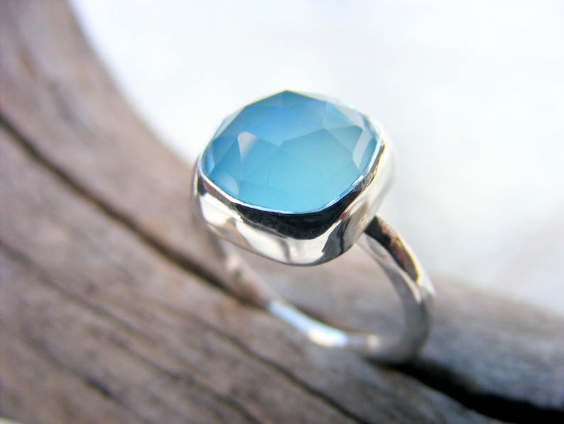 Puede incluir: Anillo de plata con una gema grande, de corte cuadrado, de color azul claro. La gema est&aacute; engastada en un bisel liso y pulido. La banda del anillo tiene un dise&ntilde;o org&aacute;nico texturizado.