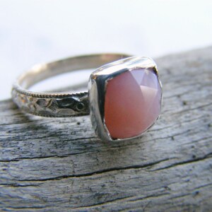 May include: A silver ring with a pink opal gemstone set in a square bezel. The ring band is decorated with a delicate floral pattern.