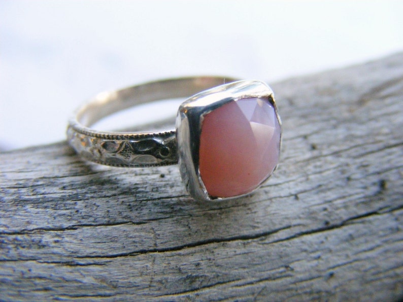 May include: A silver ring with a pink opal gemstone set in a square bezel. The ring band is decorated with a delicate floral pattern.