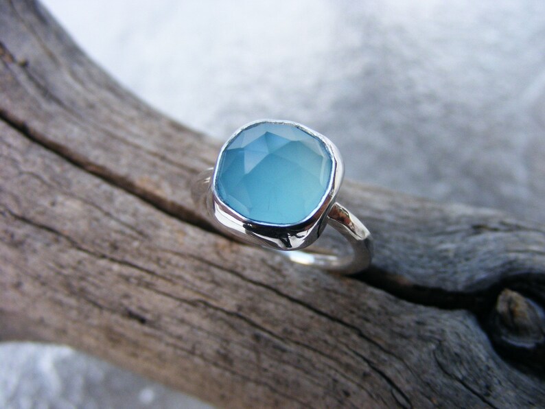 Puede incluir: Un anillo de plata con una gema grande, de corte cuadrado, de color azul claro. El anillo est&aacute; colocado sobre un trozo de madera flotante, mostrando el dise&ntilde;o y el color de la joya. La gema tiene una superficie facetada.