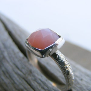 May include: A silver ring with a pink gemstone set in a bezel setting. The band is decorated with a floral design.