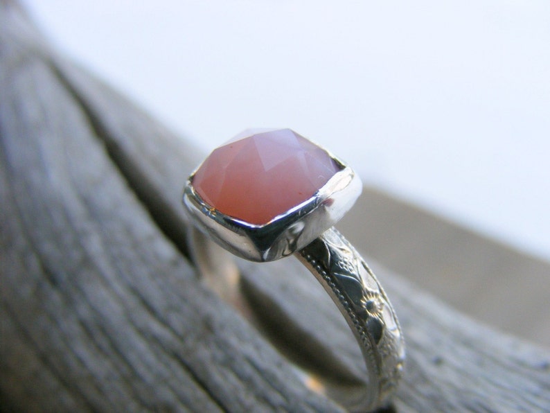 May include: A silver ring with a pink gemstone set in a bezel setting. The band is decorated with a floral design.