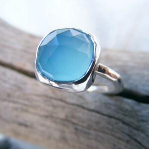 Puede incluir: Un anillo de plata con una piedra preciosa azul claro de corte cuadrado grande. La piedra preciosa tiene m&uacute;ltiples facetas, que reflejan la luz. La banda del anillo es lisa, contrastando con el engaste texturizado de la piedra.