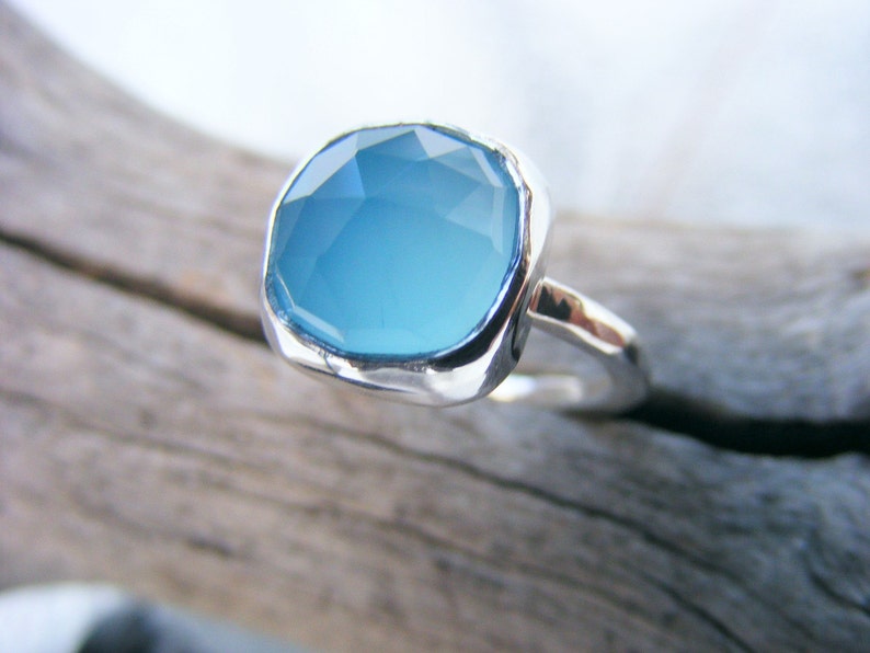 Puede incluir: Un anillo de plata con una piedra preciosa azul claro de corte cuadrado grande. La piedra preciosa tiene m&uacute;ltiples facetas, que reflejan la luz. La banda del anillo es lisa, contrastando con el engaste texturizado de la piedra.