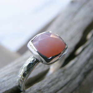 May include: A silver ring with a pink gemstone set in a square bezel. The ring band is decorated with an intricate floral design.