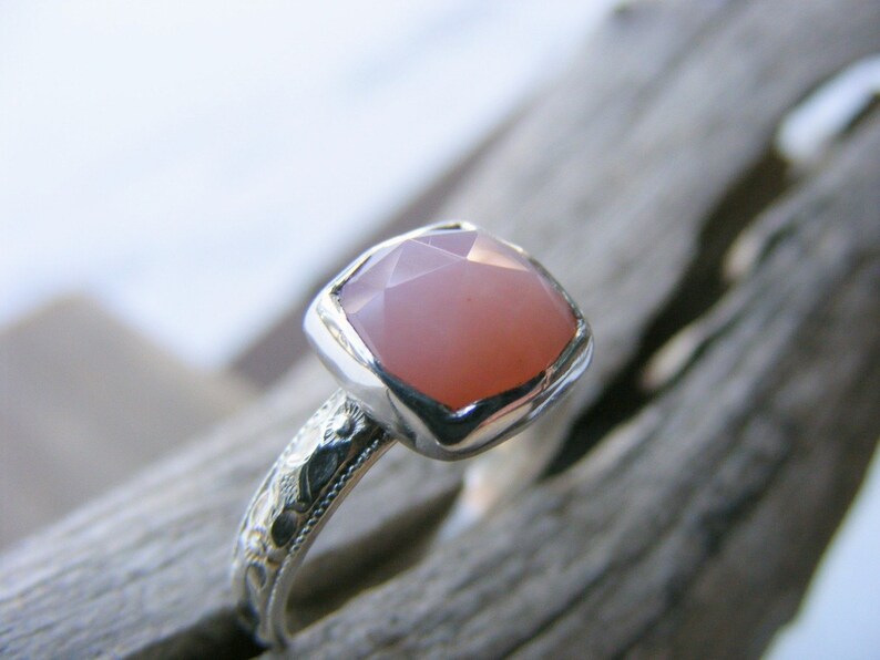 May include: A silver ring with a pink gemstone set in a square bezel. The ring band is decorated with an intricate floral design.