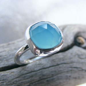 Puede incluir: Un anillo de plata con una gran piedra preciosa azul claro de talla cuadrada. La banda del anillo tiene un aspecto texturizado y martillado, y la piedra preciosa est&aacute; engastada en un bisel liso. El anillo se muestra sobre un trozo de madera flotante.