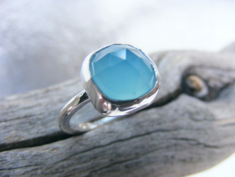 Puede incluir: Un anillo de plata con una gran piedra preciosa azul claro de talla cuadrada. La banda del anillo tiene un aspecto texturizado y martillado, y la piedra preciosa est&aacute; engastada en un bisel liso. El anillo se muestra sobre un trozo de madera flotante.