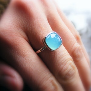 Puede incluir: Un anillo de plata con una piedra preciosa azul claro de corte cuadrado y grande. La piedra preciosa tiene m&uacute;ltiples facetas, creando un efecto brillante. El anillo tiene una banda de plata pulida y sencilla. El anillo se muestra en un dedo.