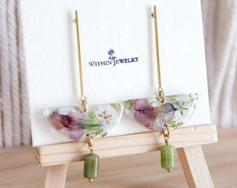 May include: A pair of earrings with a floral design. The earrings feature a clear, half-moon shape with pressed purple and white flowers, gold accents, and a green stone bead. The earrings hang from a gold bar. The brand name "WITHIN JEWELRY" is printed on the card.