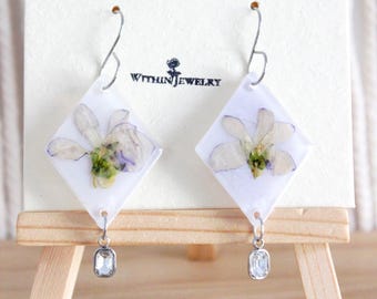 May include: A pair of handmade earrings featuring pressed flowers encased in clear resin, set in white diamond-shaped frames. Each earring has a silver hook and a small, rectangular, clear crystal charm. The earrings are displayed on a white card with the text "WITHIN JEWELRY".