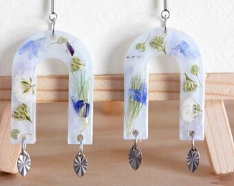 May include: A pair of unique earrings featuring a U-shaped design with embedded dried flowers in clear resin. The earrings have silver-colored metal posts and decorative charms. The brand name "WITHIN JEWELRY" is visible in the background.
