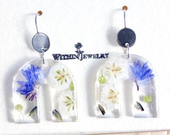 May include: Clear resin earrings with a U-shaped design, featuring blue and white flowers and green accents. The earrings have silver-colored metal hooks and a small, round top. The text "Within Jewelry" is visible.