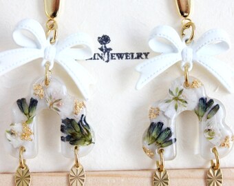 May include: A pair of elegant earrings featuring a clear, arched design filled with dried flowers and gold flakes. Each earring has a white bow at the top and a gold-colored oval charm at the bottom. The earrings are displayed on a light wooden surface.