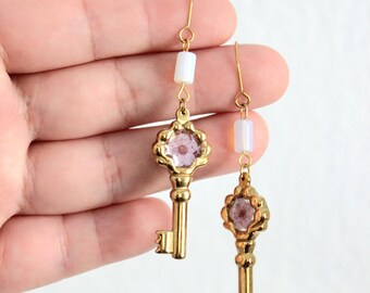 May include: A pair of gold-toned key-shaped drop earrings featuring a small white cylindrical bead at the top and a circular setting containing a light purple floral element at the center of each key head, suspended from gold-toned ear wires.