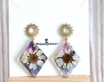 May include: A pair of diamond-shaped resin earrings featuring embedded white daisy flowers, purple petals, and gold flakes. Each earring hangs from a circular, sunburst-patterned brass stud. The jewelry is displayed on a white card labeled Within Jewelry.