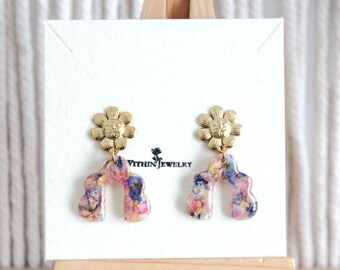 May include: A pair of earrings featuring gold-toned flower studs with dangling arch-shaped pendants. The pendants are translucent with embedded pink and blue floral elements and gold flakes. The text VITHIN JEWELRY is printed on the white display card.