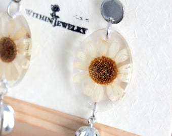 May include: Dangle earrings featuring oval clear resin pendants with embedded pressed white daisy flowers. Each earring has a silver-toned metal hook, a small circular silver disc, and a ribbed silver-toned bead at the base. The text Within Jewelry is printed on the white display card.