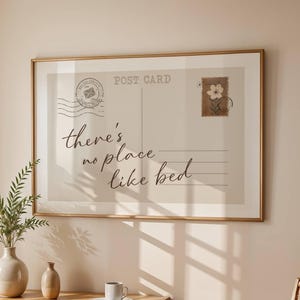 May include: A framed print resembling a postcard, with the text "there's no place like bed" in elegant script. The print has a beige background with a postage stamp graphic and is displayed above a wooden desk with a vase and greenery.