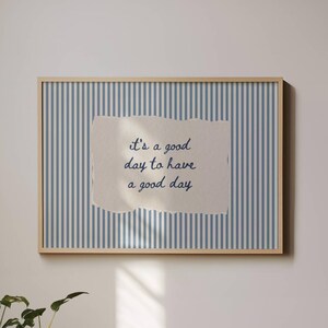 May include: A rectangular wall art print in a light wood frame featuring a blue and white vertical striped background with a central torn paper effect containing the dark blue cursive text, it's a good day to have a good day.