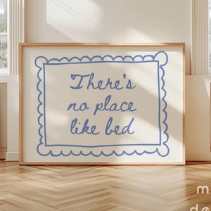 Puede incluir: Un cartel enmarcado con el texto "There's no place like bed" en azul. El texto está dentro de un borde azul festoneado sobre un fondo crema. El marco es de color marrón claro. El cartel se muestra sobre un suelo de madera.