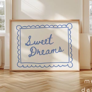 May include: A framed print with the words "Sweet Dreams" in blue script. The text is inside a blue scalloped border on a cream background. The frame is light brown. The print is displayed on a wooden floor.