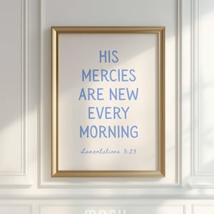 May include: A framed print with the text "HIS MERCIES ARE NEW EVERY MORNING" in light blue. The frame is gold, and the background is a cream color. The text "Lamentations 3:23" is below the main text. The print is hanging on a white wall.