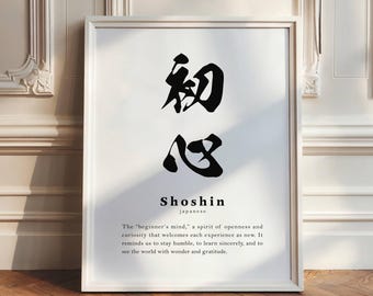 May include: A framed print with the Japanese word "Shoshin" in black calligraphy. Below the word is the English translation and a definition. The print is in a white frame and leans against a white wall.
