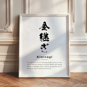 May include: White framed art print with Japanese calligraphy and the word "Kintsugi". The text below describes the art of mending broken pottery with urushi lacquer and gold, embracing imperfection. The print is leaning against a white wall on a wooden floor.