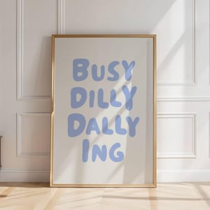 May include: A framed art print with the words "BUSY DILLY DALLY ING" in light blue, displayed against a white wall. The room features a wooden floor, a small wooden cabinet, and a vase with greenery. Natural light streams in from a window.