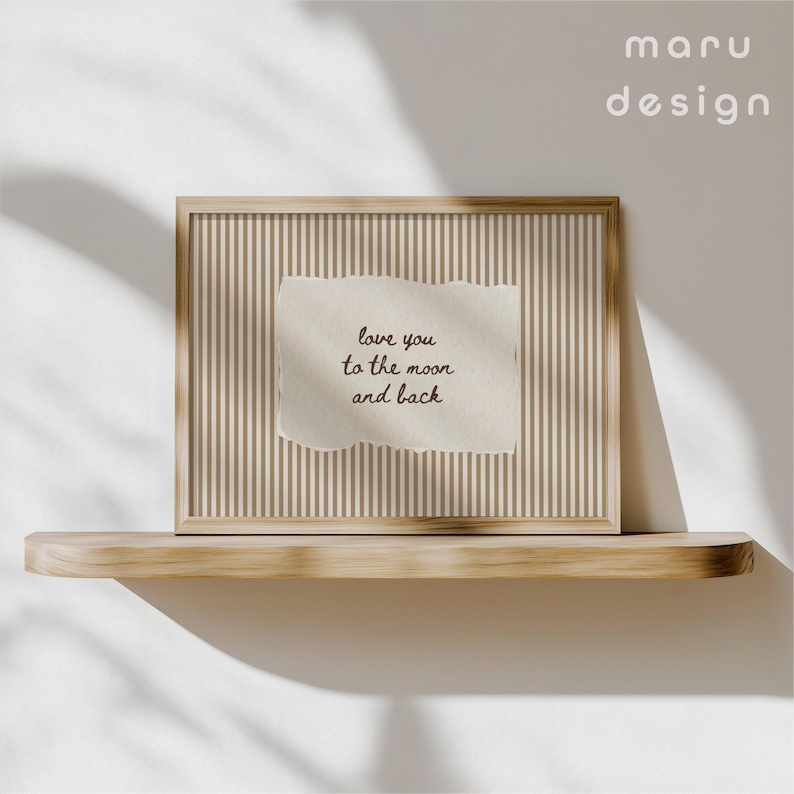 May include: A framed print with a light brown wooden frame. The print features a beige background with vertical stripes and a torn paper with the handwritten text "love you to the moon and back". The print is displayed on a wooden shelf.