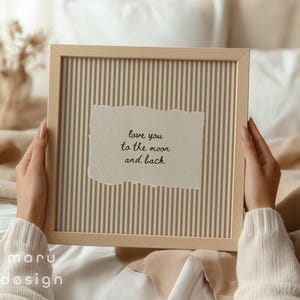 May include: A framed print with a light wood frame. The print has a striped background and a torn paper element with the text "love you to the moon and back". The frame is held by a person wearing a cream-colored sweater.