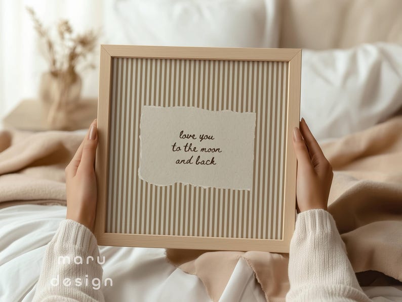 May include: A framed print with a light wood frame. The print has a striped background and a torn paper element with the text "love you to the moon and back". The frame is held by a person wearing a cream-colored sweater.