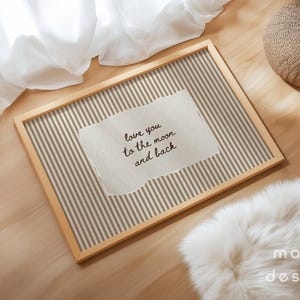 May include: A rectangular wooden frame with a striped background and a torn paper insert. The paper reads "love you to the moon and back" in black script. The frame is on a wooden surface with a white fur rug and a woven pouf.