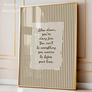 Peut inclure: Une affiche encadrée avec un cadre doré. L'œuvre présente un fond à rayures beiges et blanches avec un élément de papier déchiré au centre. Le texte sur le papier se lit : "Slow down, you're doing fine. You can't be everything you wanna be before your time."
