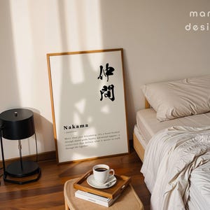 May include: A rectangular wall art print in a wooden frame featuring the Japanese kanji for Nakama, the word Nakama, and a definition text. The print is displayed on a floor next to a black lamp and a bed with beige bedding and a wooden tray with a cup.