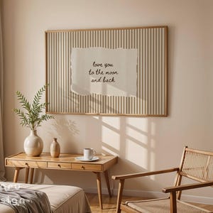 May include: A framed artwork with a vertical striped background and a torn paper element. The paper has the text "love you to the moon and back" in black script. The room also features a wooden desk, chair, and decorative vases.