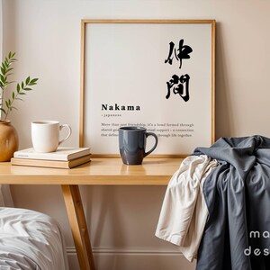 May include: A framed wall art print featuring the Japanese kanji for Nakama, the word Nakama, and a definition of the term. The print is displayed on a wooden desk alongside a white mug, a dark gray mug, a stack of books, and a green plant.