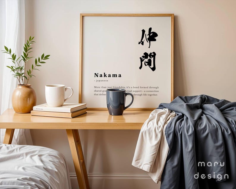 May include: A framed wall art print featuring the Japanese kanji for Nakama, the word Nakama, and a definition of the term. The print is displayed on a wooden desk alongside a white mug, a dark gray mug, a stack of books, and a green plant.