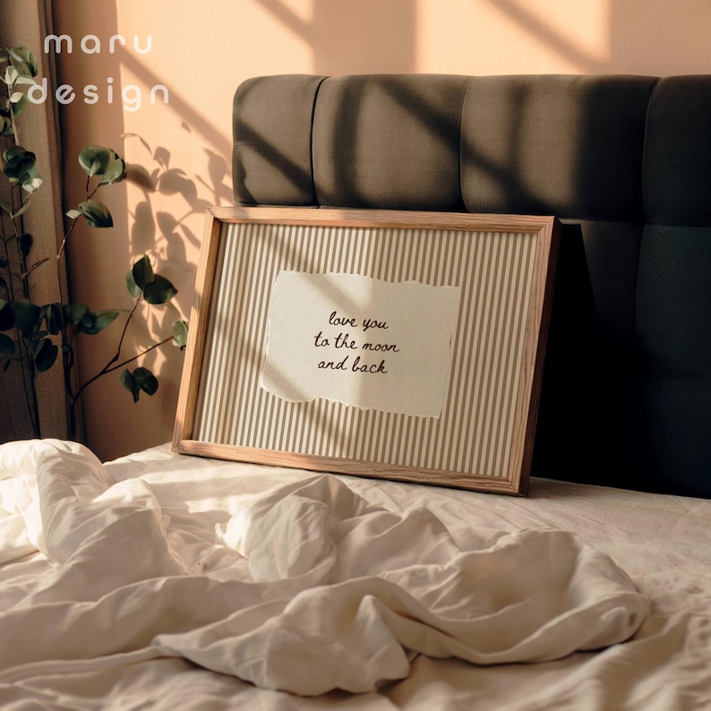 May include: A framed print with a light wood frame and a striped background. The print features a torn piece of paper with the handwritten text "love you to the moon and back". The print is on a bed with white sheets.