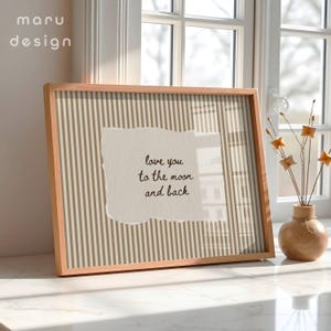 May include: A framed print with a light brown wooden frame. The print features a beige and gold striped background with a torn paper overlay. The text on the paper reads "love you to the moon and back". A small vase with decorative stems sits nearby.