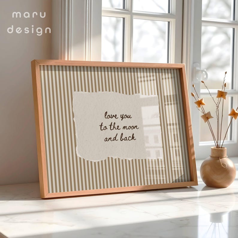 May include: A framed print with a light brown wooden frame. The print features a beige and gold striped background with a torn paper overlay. The text on the paper reads "love you to the moon and back". A small vase with decorative stems sits nearby.