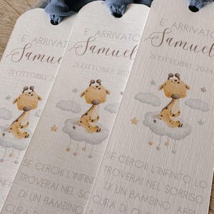 May include: Three off-white rectangular bookmarks with rounded tops and blue fabric tassels. Each features a watercolor illustration of a giraffe sitting on a cloud with stars, the text E' ARRIVATO SAMUEL 21 OTTOBRE 2024, and Italian text.
