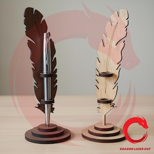 Puede incluir: Dos portalápices de madera con forma de pluma. Uno es marrón oscuro con un bolígrafo plateado, el otro es marrón claro. Cada uno tiene una base redonda y un soporte para un bolígrafo. El portalápices es de madera.