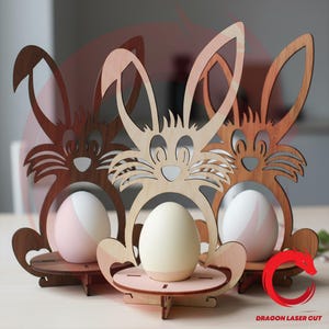 May include: Wooden Easter bunny egg holders in light and dark brown. Each bunny has a cutout face and holds a painted egg. The eggs are white, pink, and cream-colored. The design is laser cut with a Dragon Laser Cut logo.