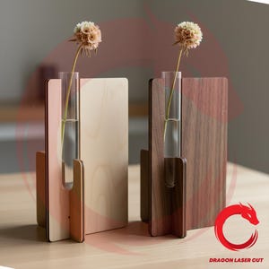 May include: Two wooden bud vases, one light and one dark, each holding a single flower in a glass tube. The vases have a modern, minimalist design with a rectangular back and a curved base. The logo "DRAGON LASER CUT" is visible.