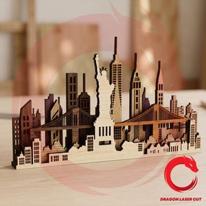 Peut inclure: Un modèle en bois découpé au laser représentant la ligne d'horizon de New York avec la Statue de la Liberté, des gratte-ciel et des ponts dans des tons de bois brun. La base porte le texte Dragon Laser Cut avec un logo de dragon rouge.