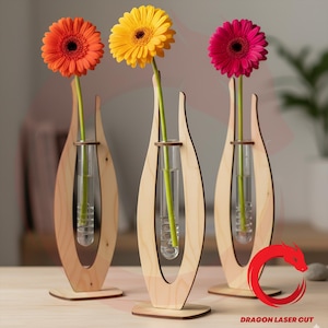 Op de afbeelding: Drie houten vazen, elk met een enkele gerbera. De bloemen zijn oranje, geel en roze. De vazen hebben een open ontwerp met een glazen buis voor de bloemstengel. De vazen staan op een houten oppervlak.